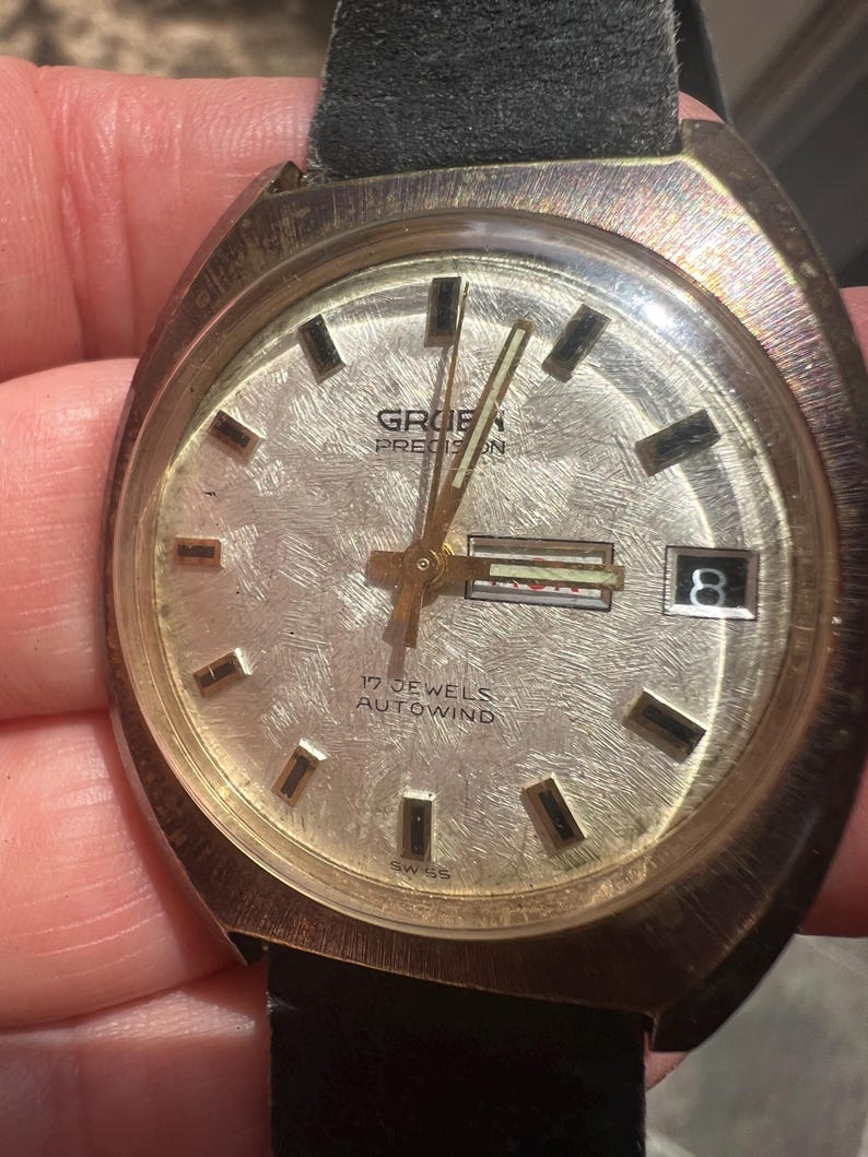 Op de afbeelding: Vintage Gruen Precision polshorloge met een getextureerde zilveren wijzerplaat, goudkleurige wijzers en markeringen, en een datumvenster. Het horloge heeft een zwarte leren band en de tekst "17 JEWELS AUTOWIND" is zichtbaar.