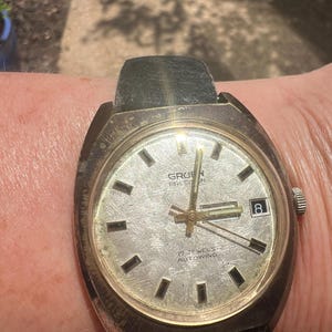 Op de afbeelding: Vintage Gruen Precision horloge met een zilveren wijzerplaat, zwarte rechthoekige uurmarkeringen en een datumvenster op 3 uur. Het horloge heeft een goudkleurige kast en een zwarte leren band. De tekst "17 JEWELS AUTOWIND" is zichtbaar.