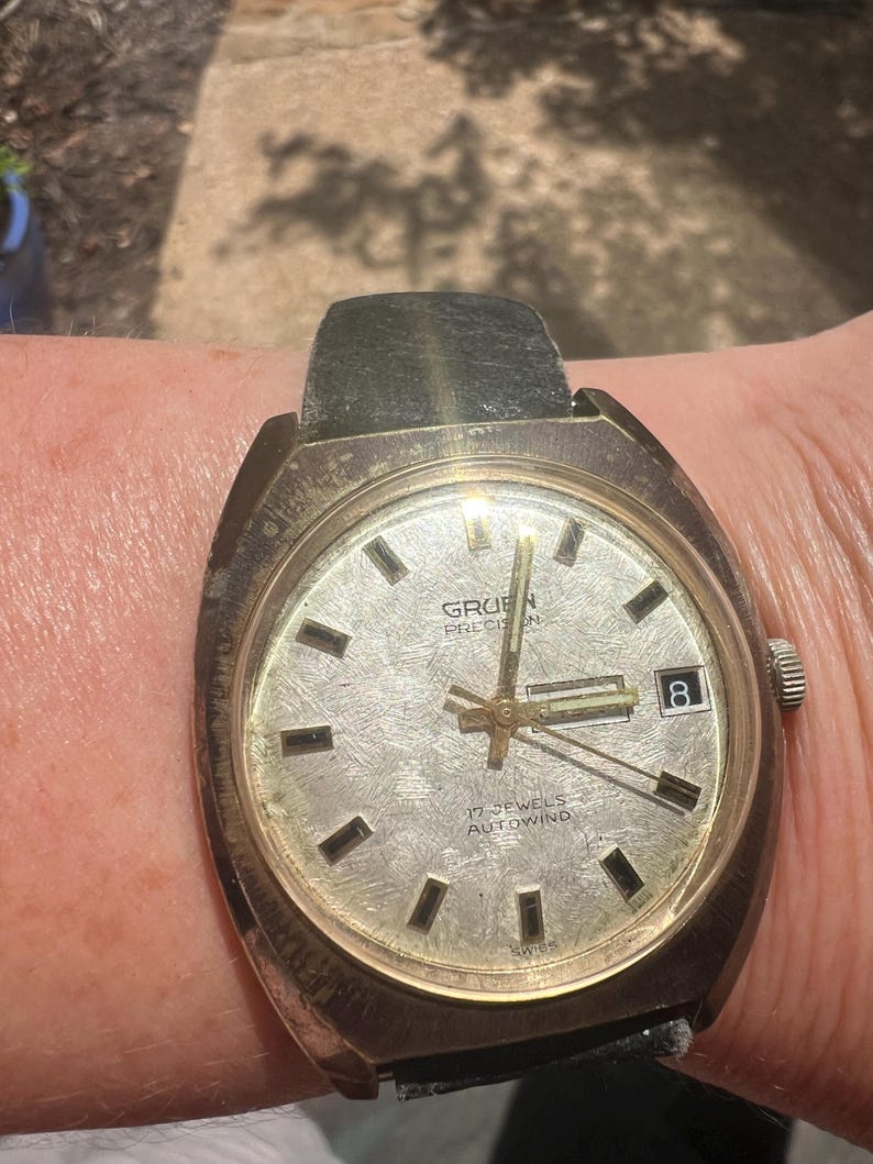 Op de afbeelding: Vintage Gruen Precision horloge met een zilveren wijzerplaat, zwarte rechthoekige uurmarkeringen en een datumvenster op 3 uur. Het horloge heeft een goudkleurige kast en een zwarte leren band. De tekst "17 JEWELS AUTOWIND" is zichtbaar.