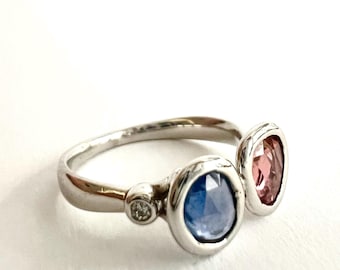 May include: A silver ring featuring a blue gemstone, a pink gemstone, and a small diamond. The ring has a modern design with the gemstones set in bezel settings. The ring is displayed on a white background.