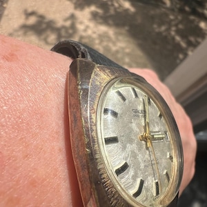 Op de afbeelding: Vintage Gruen Precision polshorloge met een zilverkleurige wijzerplaat, gouden wijzers en zwarte markeringen. Het horloge heeft een zwarte leren band en is gemarkeerd met "17 Jewels Autowind". Het horloge wordt om de pols gedragen.