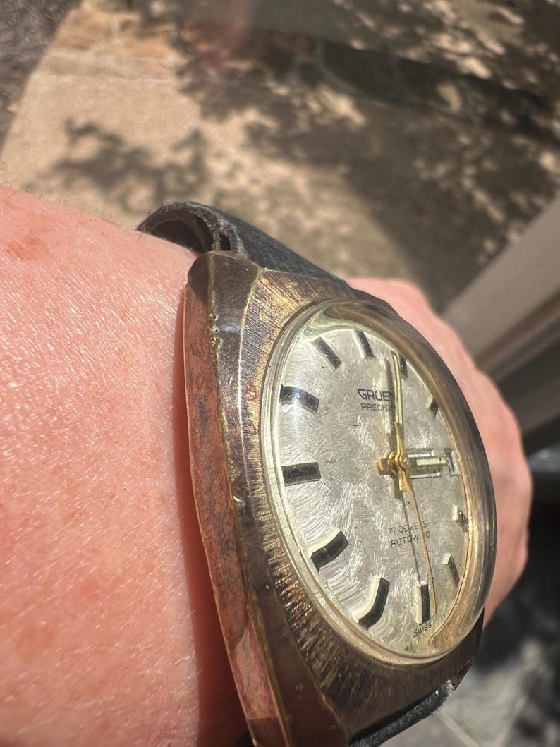 Op de afbeelding: Vintage Gruen Precision polshorloge met een zilverkleurige wijzerplaat, gouden wijzers en zwarte markeringen. Het horloge heeft een zwarte leren band en is gemarkeerd met "17 Jewels Autowind". Het horloge wordt om de pols gedragen.