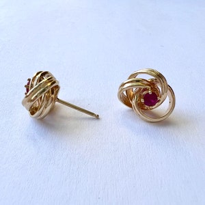 May include: A pair of gold earrings with a red gemstone in the center of each earring. The earrings are designed in a knot shape.