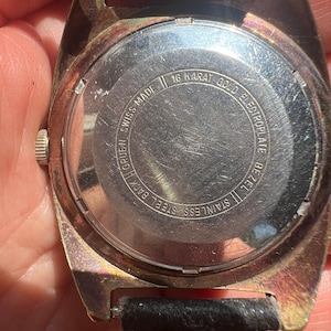 Op de afbeelding: Close-up van een vintage Gruen-horloge. Het horloge heeft een goudkleurige lunette en een zwarte leren band. De achterkant van het horloge is van roestvrij staal met de tekst "16 Karat Gold Electroplate Bezel" en "Swiss Made".