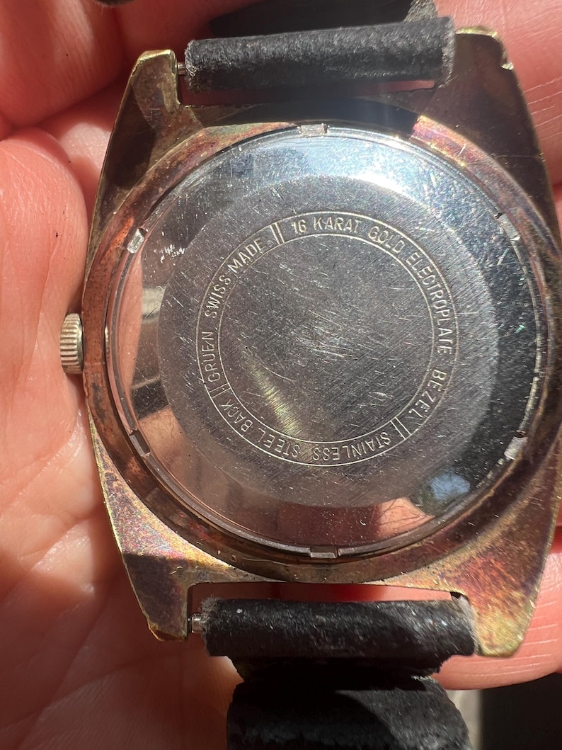 Op de afbeelding: Close-up van een vintage Gruen-horloge. Het horloge heeft een goudkleurige lunette en een zwarte leren band. De achterkant van het horloge is van roestvrij staal met de tekst "16 Karat Gold Electroplate Bezel" en "Swiss Made".