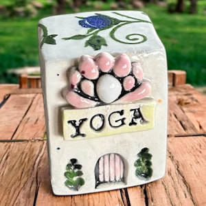 May include: A rectangular ceramic plaque with a white base, featuring a pink lotus flower design and the word "YOGA" in black. The plaque has a small arched doorway and floral accents. The background is blurred, suggesting an outdoor setting.