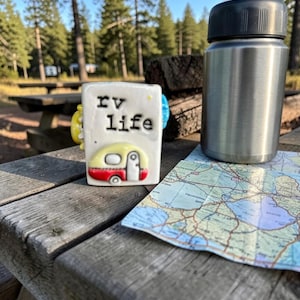 Könnte beinhalten: Eine Keramik-Tasse mit dem Text "RV LIFE" und einem Cartoon-Wohnwagen-Design, zusammen mit einer Edelstahl-Wasserflasche und einer Karte auf einem Picknicktisch aus Holz. Die Tasse hat gelbe, rote und blaue Akzente.