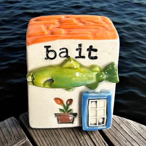 May include: A ceramic bait box with a textured orange roof and the word "bait" in black letters. A green fish is centered on the front, with a small blue window and a potted plant below. The box is on a wooden dock.