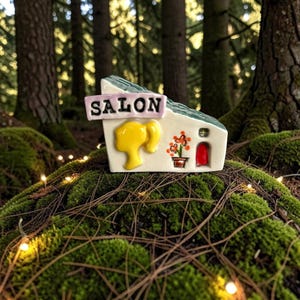 May include: A whimsical ceramic miniature salon with a green roof and the word "SALON" on a sign. Features a yellow hair design, a red door, and a flower pot. Set in a mossy forest with string lights.