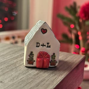 May include: A small, white ceramic house with a gray roof and a red heart above the letters "D + L". The house has a red door and two potted plants on either side. The house is sitting on a wooden surface.
