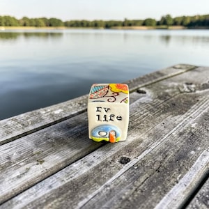 Könnte beinhalten: Kleine, rechteckige Keramikfigur mit dem Aufdruck "RV life". Die Figur zeigt ein farbenfrohes Wohnwagen-Design und steht auf einem verwitterten Holzsteg mit See und Bäumen im Hintergrund.