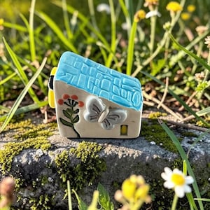 May include: A small ceramic house figurine featuring a textured light blue roof, a white wall with a raised grey butterfly relief, a painted orange flower, and a small yellow door. The structure has a flat base and a slanted roof design.