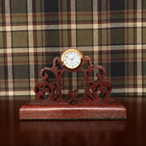 May include: A small, round, white-faced clock with gold-coloured hands and numbers is mounted on a dark red wooden stand with intricate cutouts. The stand has a rectangular base.