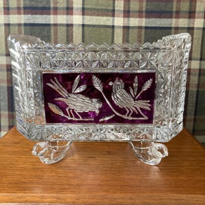 May include: A clear glass bowl with a purple, etched design of two birds and wheat stalks. The bowl has a rectangular shape and sits on three feet.
