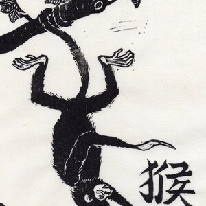May include: Black and white woodblock print of a monkey hanging upside down from a tree branch. The monkey is in the center of the image and the tree branch is above it. The monkey is facing the viewer and has its arms and legs outstretched. The word "猴" is written in black ink below the monkey.