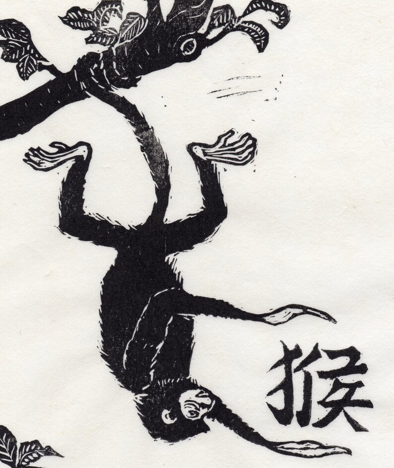 May include: Black and white woodblock print of a monkey hanging upside down from a tree branch. The monkey is in the center of the image and the tree branch is above it. The monkey is facing the viewer and has its arms and legs outstretched. The word "猴" is written in black ink below the monkey.