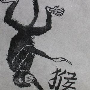 May include: Black and white woodcut print of a monkey hanging upside down with the Chinese character for "monkey" printed below.