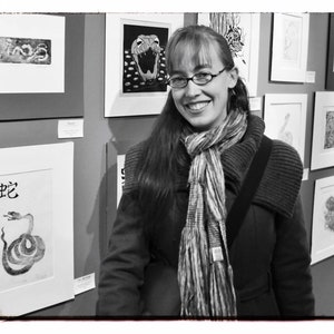 May include: A woman with long brown hair wearing a black coat and a striped scarf stands in front of a wall of framed black and white prints. The prints feature various designs, including snakes, animals, and abstract patterns.