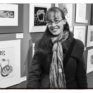 May include: A woman with long brown hair wearing a black coat and a striped scarf stands in front of a wall of framed black and white prints. The prints feature various designs, including snakes, animals, and abstract patterns.