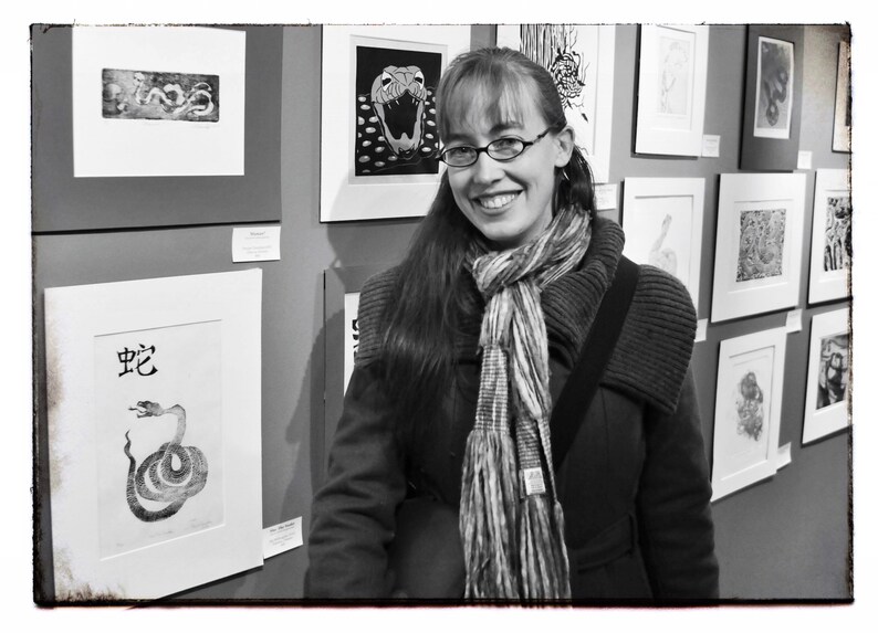 May include: A woman with long brown hair wearing a black coat and a striped scarf stands in front of a wall of framed black and white prints. The prints feature various designs, including snakes, animals, and abstract patterns.
