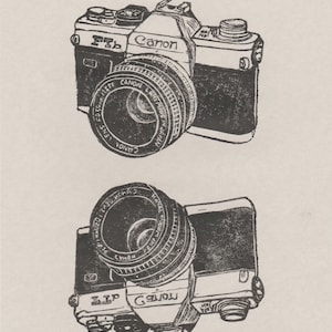 May include: A black and white illustration of a Canon FTb camera with a lens. The camera is labeled "Canon" and "FTb". The illustration is signed "Ele Willowy Way 2014" and numbered "3/10".