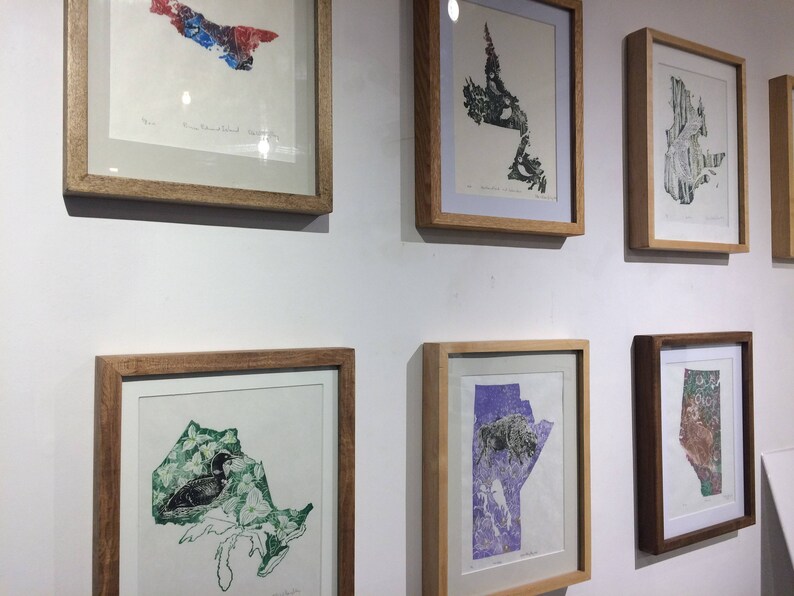 K&ouml;nnte beinhalten: Eine Wandmontage von gerahmten Kunstwerken mit verschiedenen Karten kanadischer Provinzen. Die Karten sind in verschiedenen Farben und Stilen gedruckt, darunter eine Karte von Ontario mit einem Seetaucher, eine Karte von Manitoba mit einem Bison und eine Karte von Prince Edward Island mit einem Blumendesign.