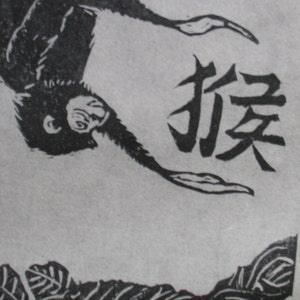 May include: Black and white woodblock print of a monkey hanging upside down with the Japanese character for "monkey" written in black ink.
