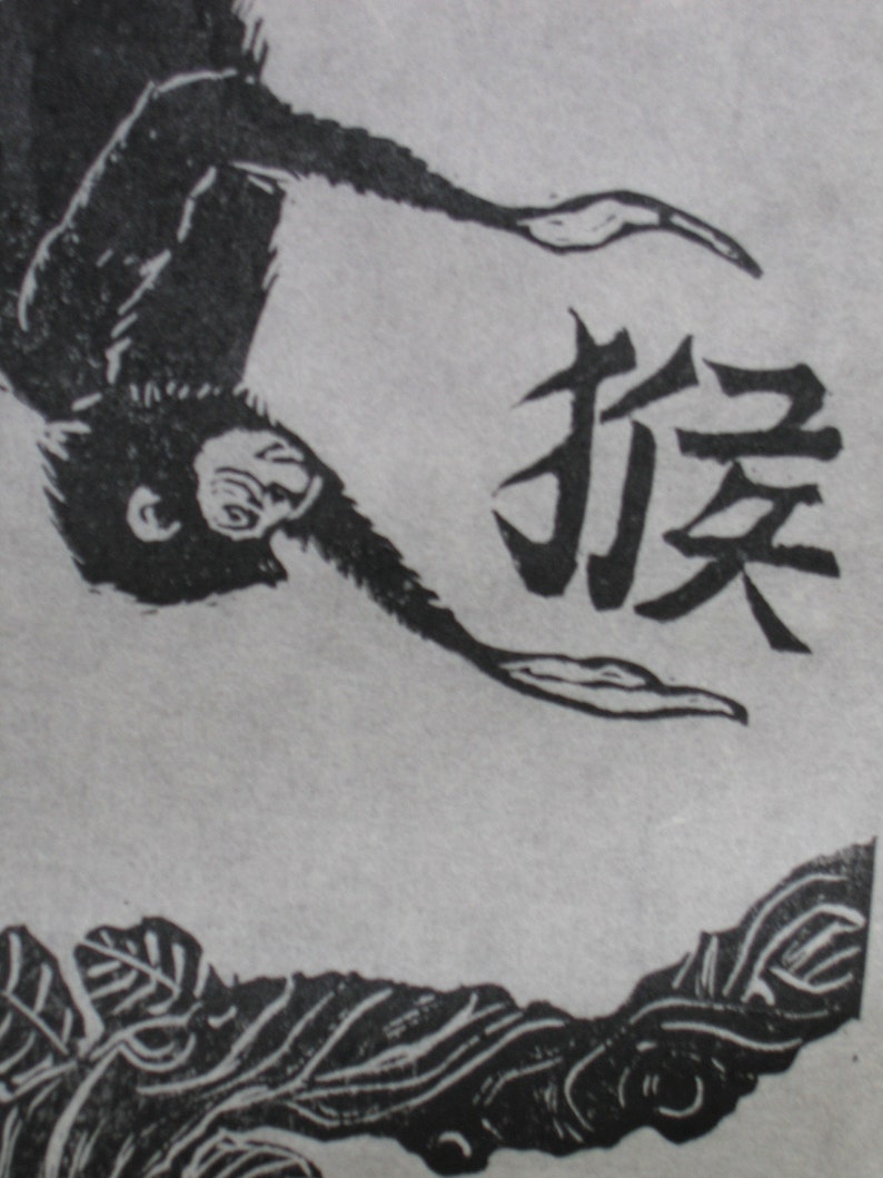 May include: Black and white woodblock print of a monkey hanging upside down with the Japanese character for "monkey" written in black ink.