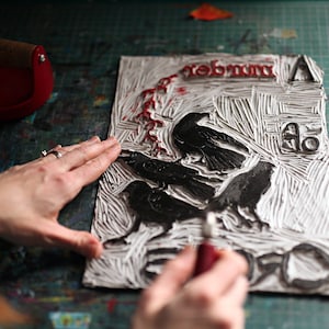 May include: A close-up of a white linoleum block print with black ink. The print features three crows and the word "remember" in red ink. A person's hand is using a tool to carve the block.