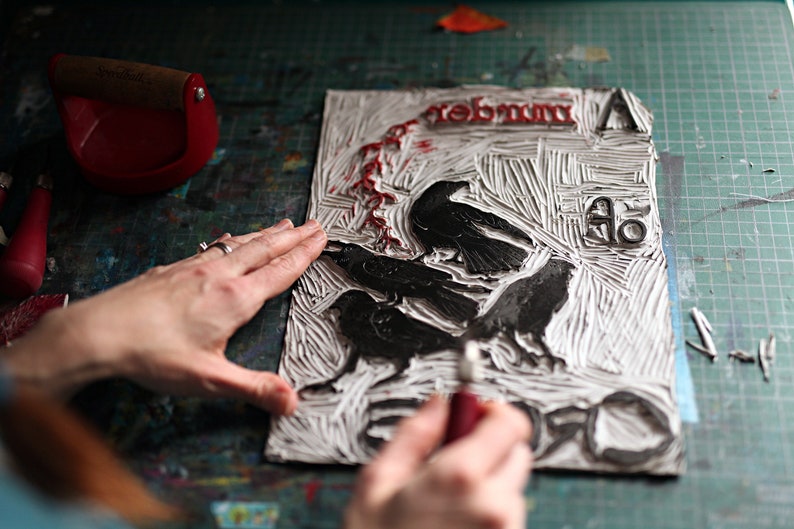 May include: A close-up of a white linoleum block print with black ink. The print features three crows and the word "remember" in red ink. A person's hand is using a tool to carve the block.