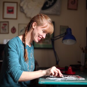 May include: A woman with long braided hair is working on a craft project at a table. She is using a tool to apply a piece of paper to a larger piece of paper. The table is covered in a green mat and there are various art supplies on the table.
