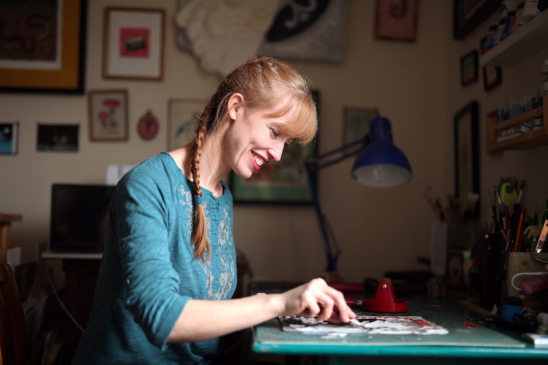 May include: A woman with long braided hair is working on a craft project at a table. She is using a tool to apply a piece of paper to a larger piece of paper. The table is covered in a green mat and there are various art supplies on the table.