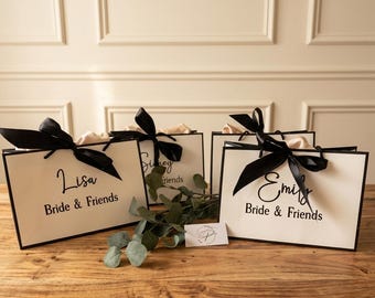 Può includere: Quattro sacchetti regalo bianchi con bordi neri e fiocchi neri, ciascuno con un nome e "Bride & Friends" in corsivo nero. Un rametto di verde e una piccola carta poggiano su una superficie di legno.