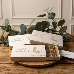 Pode incluir: Várias caixas bege de chocolates com letras douradas e um desenho de lua crescente estão dispostas sobre uma superfície de madeira. As caixas estão abertas, revelando os chocolates. Um ramo de folhas verdes é colocado perto das caixas.