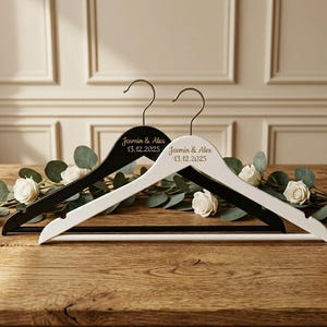May include: Two wooden clothes hangers, one black and one white, with the names "Jasmin & Alex" and the date "13.12.2025" printed on them. The hangers are resting on a wooden surface, decorated with white roses and greenery.