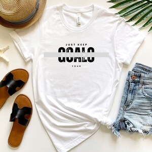 Könnte beinhalten: Weißes T-Shirt mit dem Aufdruck "Just Keep Nailing Your Goals" in Schwarz. Das Shirt liegt flach auf einer weißen Oberfläche mit einem Strohhut, Sandalen und einer Jeans-Shorts.