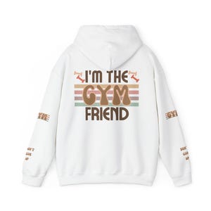 Könnte beinhalten: Weißer Kapuzenpullover mit dem Text "I'm the Gym Friend" in braunen Buchstaben mit einem Retro-Regenbogen-Design. Die Ärmel haben den Text "Don't Give Up" in braunen Buchstaben.
