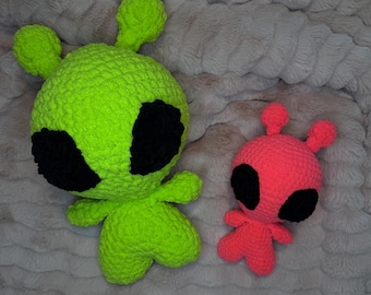 May include: Two crocheted alien plushies. The larger one is green with black eyes and the smaller one is pink with black eyes.