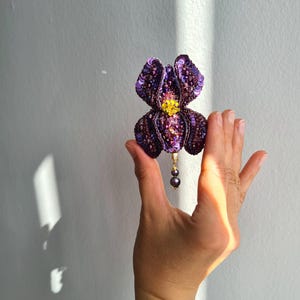 May include: A handcrafted, sequined iris brooch in shades of purple, with a yellow center and a dangling pearl accent. The floral design is detailed and dimensional, perfect for adding a touch of elegance to any outfit.