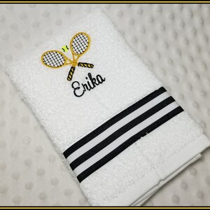 K&ouml;nnte beinhalten: Wei&szlig;es Handtuch mit gestickten Tennisschl&auml;gern und einem gelb-schwarzen Tennisball. Der Name "Erika" ist in schwarzer Schrift gestickt. Das Handtuch hat einen schwarz-wei&szlig; gestreiften Rand.