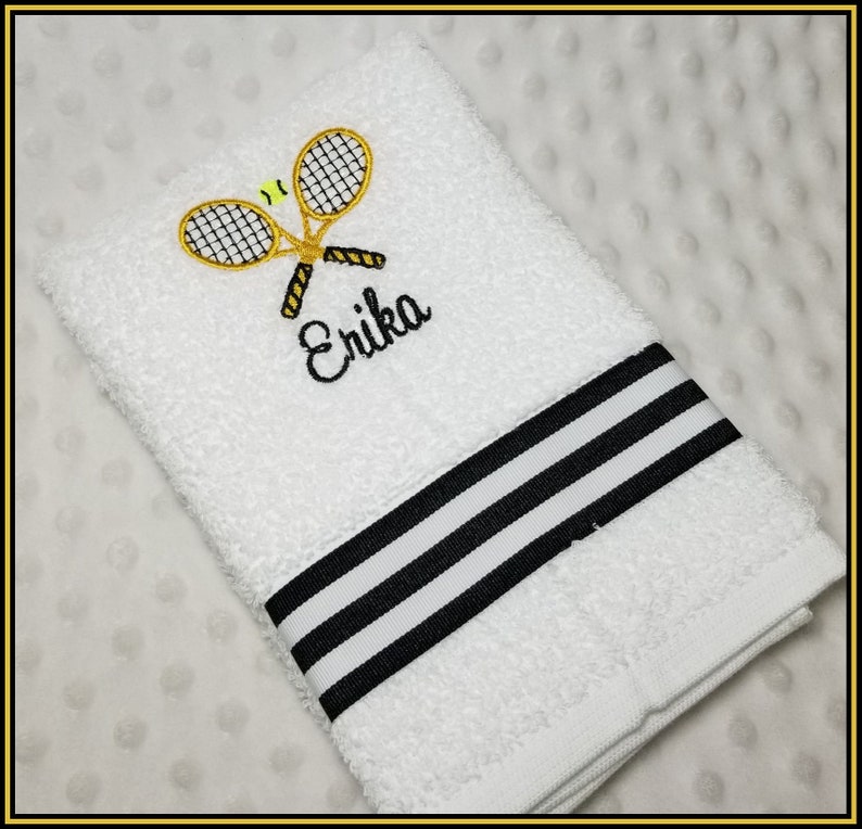 K&ouml;nnte beinhalten: Wei&szlig;es Handtuch mit gestickten Tennisschl&auml;gern und einem gelb-schwarzen Tennisball. Der Name "Erika" ist in schwarzer Schrift gestickt. Das Handtuch hat einen schwarz-wei&szlig; gestreiften Rand.