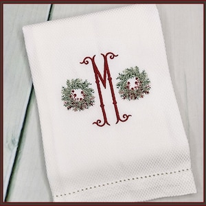 Könnte beinhalten: Ein weißes Küchentuch mit einem roten gestickten Monogramm "M" und zwei grünen Kränzen mit roten Beeren.