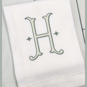 Könnte beinhalten: Weißes Handtuch mit einem gestickten Buchstaben "H" in einem dezenten Grünton. Das Handtuch hat ein strukturiertes Muster und eine genähte Bordüre. Der Buchstabe ist mittig platziert, mit sternförmigen Verzierungen auf beiden Seiten.