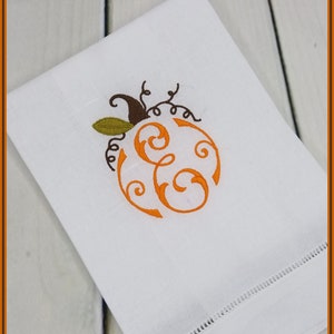 May include: A white kitchen towel with an embroidered orange pumpkin with a brown stem and green leaves. The pumpkin has a swirl design inside and the letter "E" is visible.