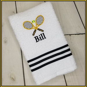 K&ouml;nnte beinhalten: Wei&szlig;es Handtuch mit gestickten Tennisschl&auml;gern, einem Tennisball und dem Namen "Bill" in Schwarz. Das Handtuch hat einen schwarz-wei&szlig; gestreiften Rand. Das Design ist auf dem Handtuch zentriert.