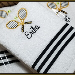 K&ouml;nnte beinhalten: Wei&szlig;es, besticktes Tennishandtuch mit schwarz-wei&szlig;em Streifenmuster. Das Handtuch zeigt ein Tennis-Schl&auml;ger- und Ball-Design mit dem Namen "Erika" in Schwarz gestickt. Die Handt&uuml;cher sind aus weichem, saugf&auml;higem Material.