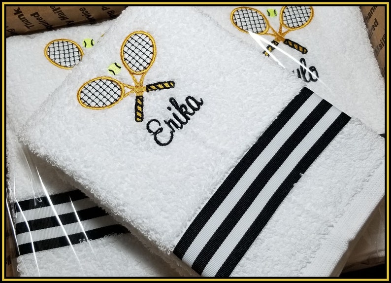 K&ouml;nnte beinhalten: Wei&szlig;es, besticktes Tennishandtuch mit schwarz-wei&szlig;em Streifenmuster. Das Handtuch zeigt ein Tennis-Schl&auml;ger- und Ball-Design mit dem Namen "Erika" in Schwarz gestickt. Die Handt&uuml;cher sind aus weichem, saugf&auml;higem Material.