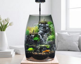 May include: A glass terrarium aquarium featuring a dark gray tinted vessel, a black overhead light, and a wooden base. Inside, a rocky landscape with green moss, miniature wooden bridges, and small figurines sits above a gravel bed with two fish.