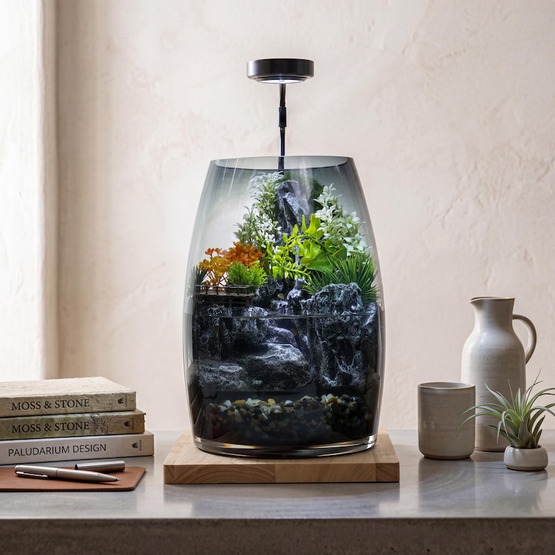 May include: A glass terrarium with a dark gradient finish containing a miniature landscape of grey rocks, green foliage, and orange plants. A black LED light fixture is mounted above the vessel, which sits on a square wooden base.