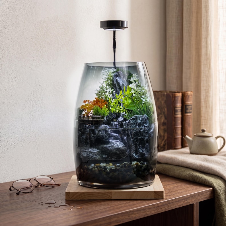 May include: A glass terrarium with a dark gradient finish containing a miniature landscape with grey rocks, green and orange artificial plants, and a small wooden platform. A black light fixture is mounted above the glass, and the base is wood.